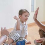 A young boy sitting on the floor high-fives a woman, with a man watching nearby