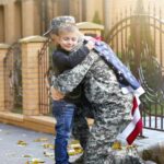 A military parent in uniform hugs their child outdoors with an American flag.