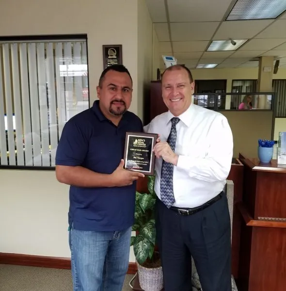 Two men standing in an office, one presenting a plaque award to the other.