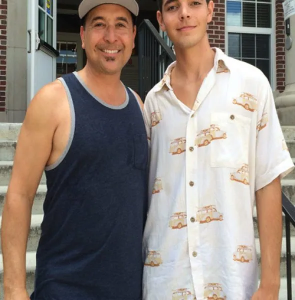 Two men standing on outdoor steps, one in a navy tank top and cap, the other in a white shirt with car prints.