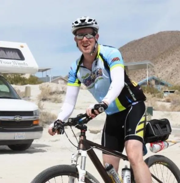 Cyclist in blue and black gear with helmet and sunglasses, posing with mountain bike outdoors.