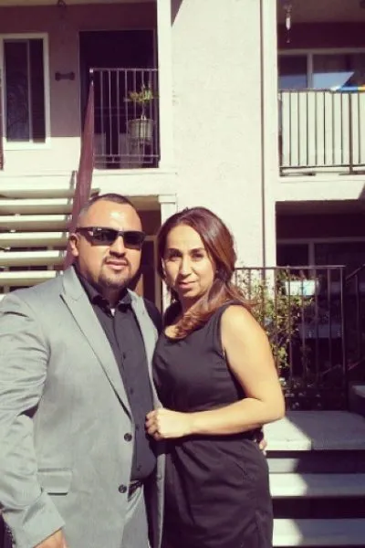 Man in gray suit and sunglasses with woman in black dress standing outdoors by residential building.