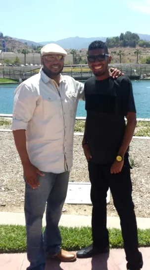 Two men standing outdoors near a body of water with hills in the background, one wearing a white shirt and cap, the other in black clothing.