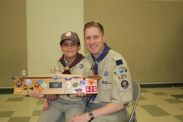 Two Boy Scouts in uniform sitting indoors, holding a wooden plaque with badges and the number 626.