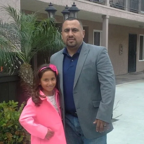 Man in gray blazer and young girl in bright pink coat posing outdoors near palm plants.