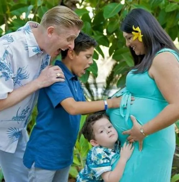 A family of four outdoors, with two boys and a man excitedly touching a pregnant woman's belly.