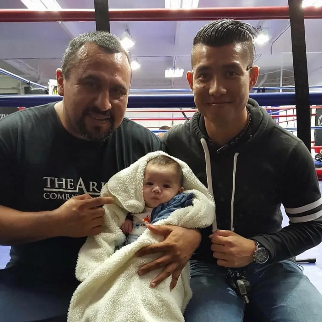 Two men sitting in a boxing ring holding a baby wrapped in a white blanket.