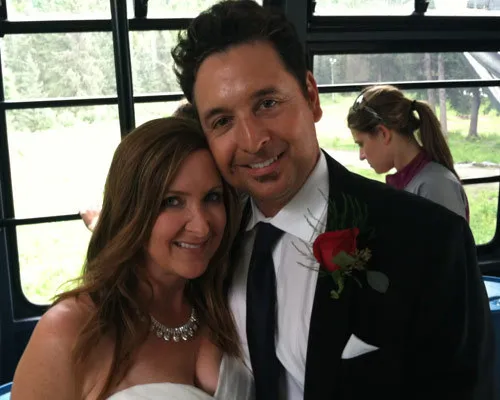 Man in a black suit with a red rose boutonniere and woman in a white dress smiling indoors.