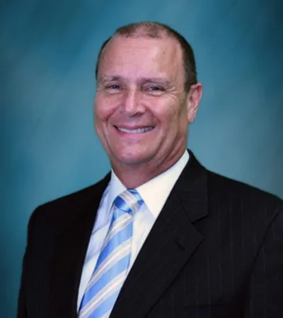 Portrait of a middle-aged man in a dark suit and blue striped tie, smiling against a blue background.