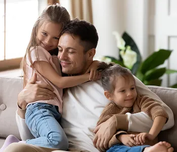 Father sitting on a beige couch warmly embracing two young daughters indoors.