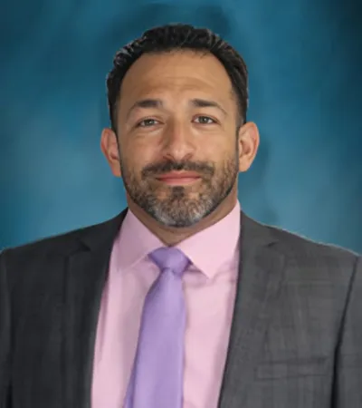 Professional headshot of a man with dark hair and beard, wearing a gray suit, pink shirt, and lavender tie.