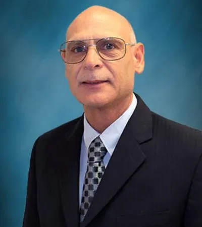 Formal portrait of a bald man wearing glasses, a dark suit, and patterned tie against a blue background.