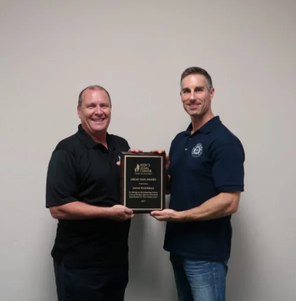 Two men wearing dark polo shirts holding a framed award plaque against a plain beige wall.