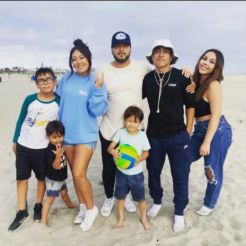 Group of seven diverse people posing on sandy beach, two children holding a volleyball.