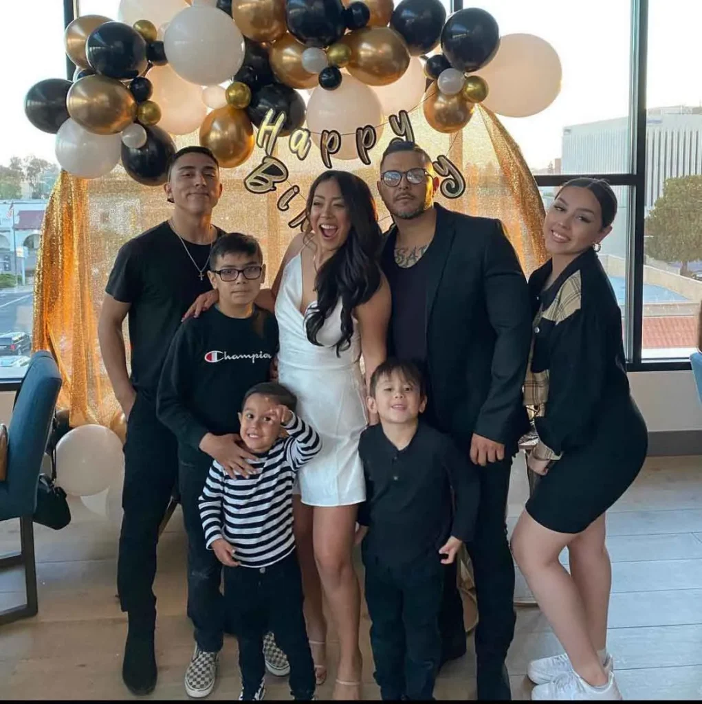 Group of seven people posing in front of gold, black, and white balloons with "Happy Birthday" banner.
