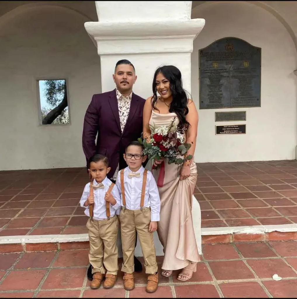 Formal family portrait with man in purple suit, woman in beige dress holding bouquet, and two boys in white shirts and tan suspenders.