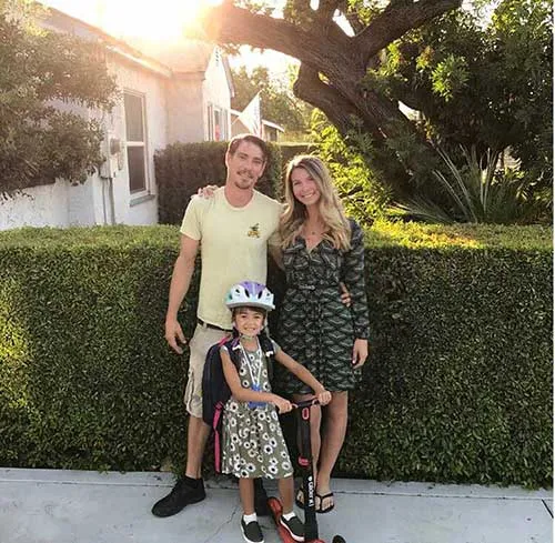 A family of three standing outdoors by a green hedge, with a child wearing a helmet on a scooter.