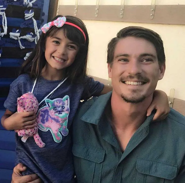 Smiling man and young girl with a pink headband and stuffed animal, embracing.