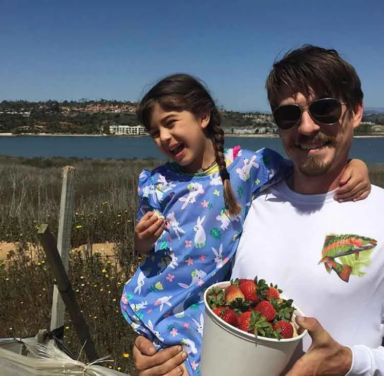 Man wearing sunglasses holding a young girl in a blue dress and a bucket of strawberries outdoors.