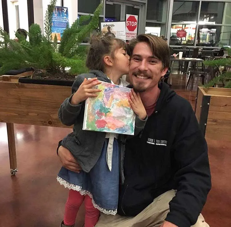 Young girl kissing a smiling man on the cheek while holding a colorful painting indoors.
