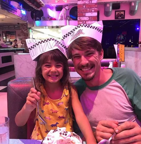 A smiling man and young girl wearing diner paper hats, seated at a booth with a dessert topped with whipped cream.