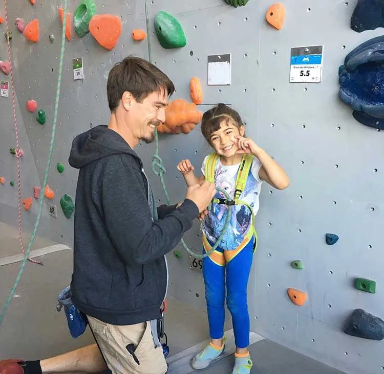 Man assisting young girl in yellow climbing harness at indoor rock climbing wall with colorful holds.