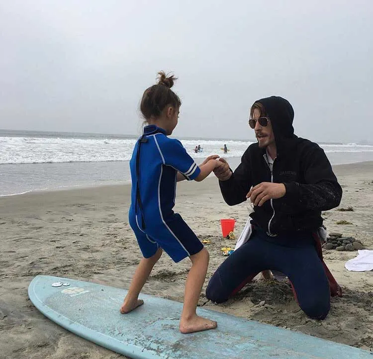 Adult in black hoodie assists child in blue wetsuit standing on surfboard at beach.
