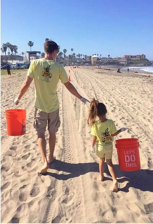 Adult and child in matching yellow shirts holding orange buckets, walking on sandy beach.