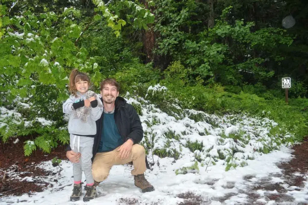 Man and young girl smiling, standing on snow-covered ground with green foliage background.