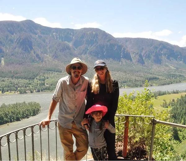 Three people posing on a balcony overlooking a river and mountainous landscape under a clear sky.
