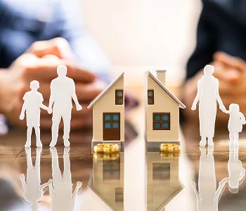 Two miniature wooden houses with gold coins and white paper cutouts of families on a reflective surface.