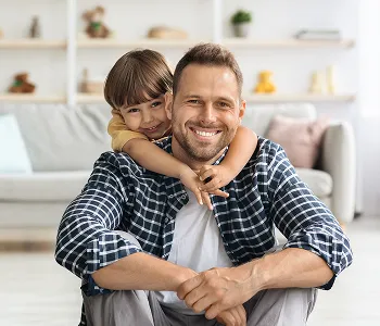 Smiling man in checkered shirt sitting with child embracing him from behind in living room.