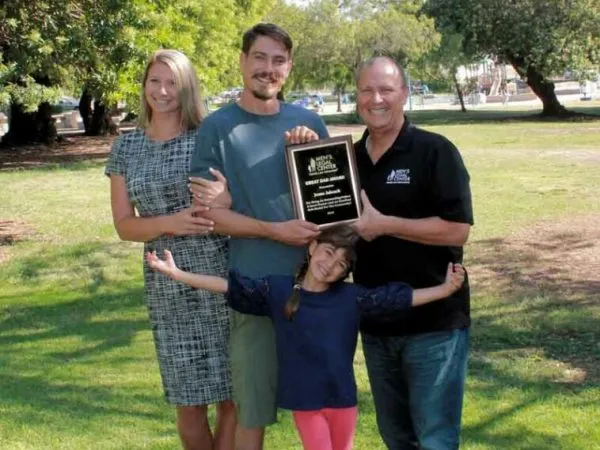 Four people standing outdoors on grass, holding a black and gold award plaque, smiling.