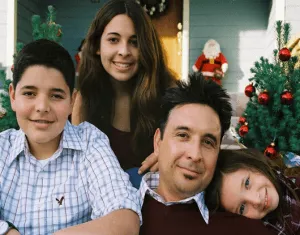 Family of four posing in front of a decorated Christmas tree and holiday ornaments.