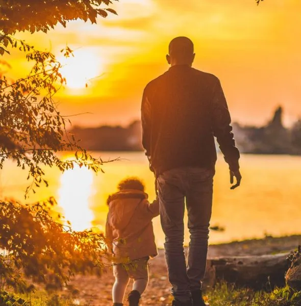 Man and child holding hands walking by a lakeside at sunset with warm orange hues.