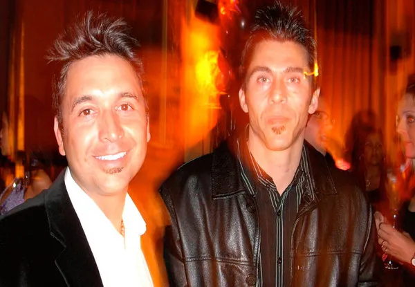 Two men dressed in dark jackets posing indoors with warm, blurred background lighting.