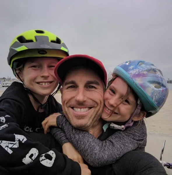 Smiling man wearing red cap hugging two children with helmets near waterfront.