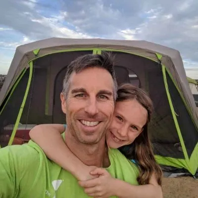 A man and young girl smiling, posing in front of a green and gray camping tent outdoors.