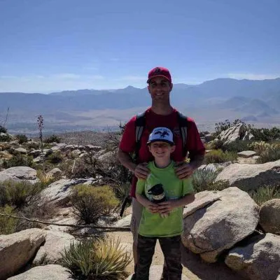 Two males, one adult and one child, standing among desert rocks with mountains in the background under a clear blue sky.
