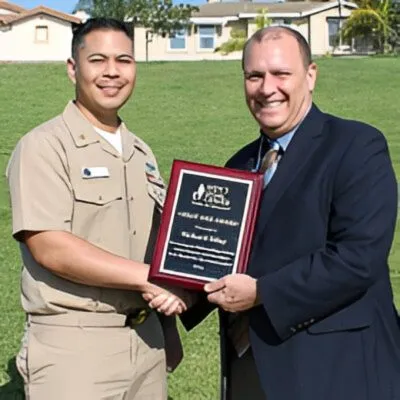 Two men smiling outdoors, one in military uniform and the other in a suit, holding a framed award plaque.