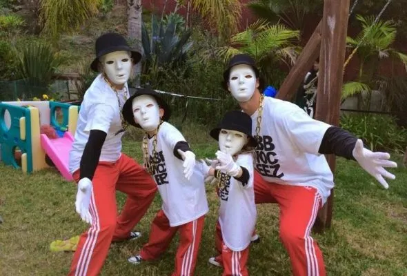 Four individuals wearing white masks, black hats, white shirts, and red pants posing outdoors on grass.