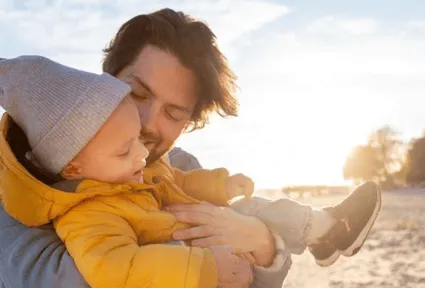 Man in gray sweater holding child in yellow jacket and gray beanie outdoors at sunset.