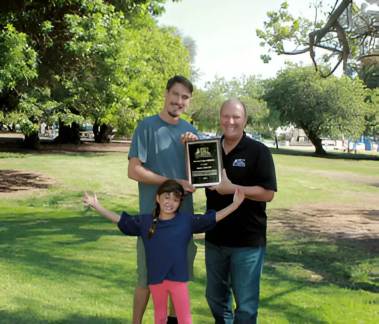 Two men holding an award plaque outdoors in a park, with a young girl in pink pants and blue shirt standing in front with arms outstretched.