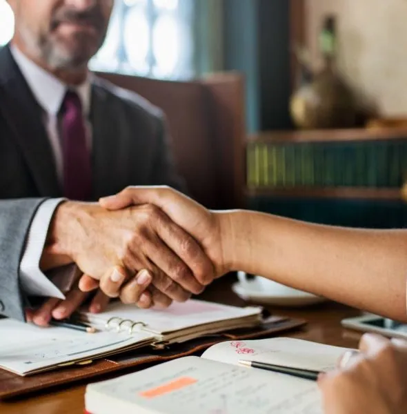 Two individuals in formal attire shaking hands over a desk with open notebooks and documents.