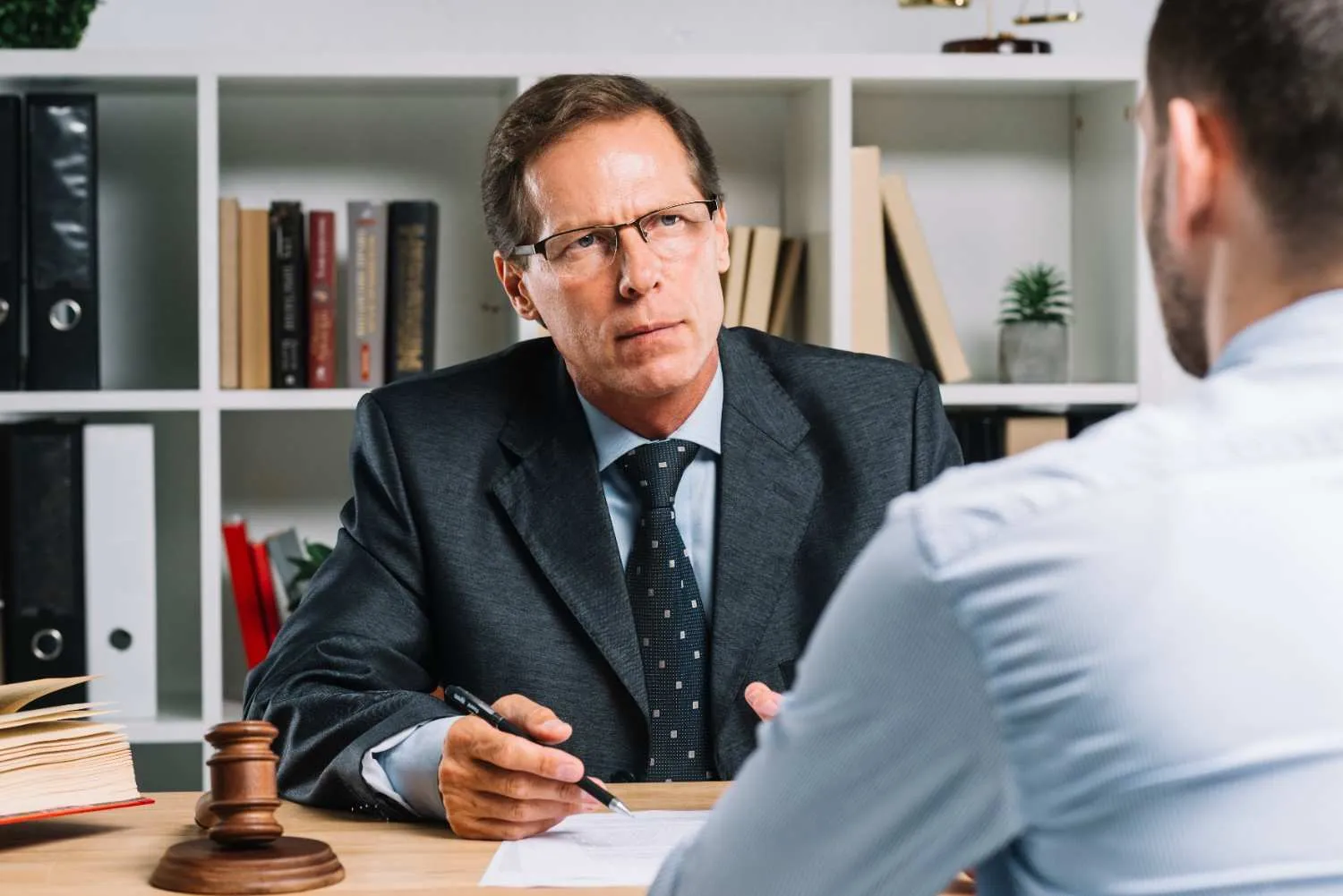Two men in a professional office setting, one in a suit holding a pen, legal books and gavel on desk.