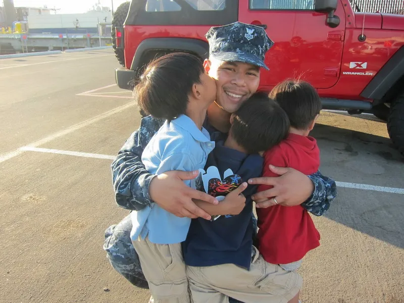 Service member in navy uniform smiling and hugging three children in a parking lot near a red Jeep Wrangler.