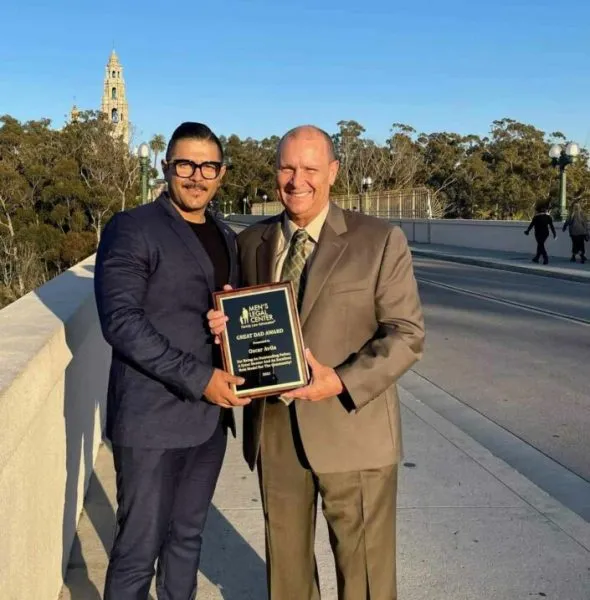 Two men in formal attire standing outdoors on a bridge, holding a plaque award.