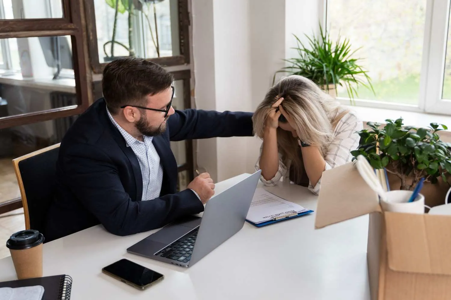 Man in suit comforting distressed woman at desk with laptop and documents.