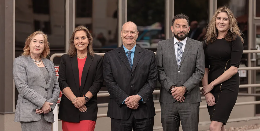 Group of five professionally dressed individuals standing outdoors in business attire, smiling.