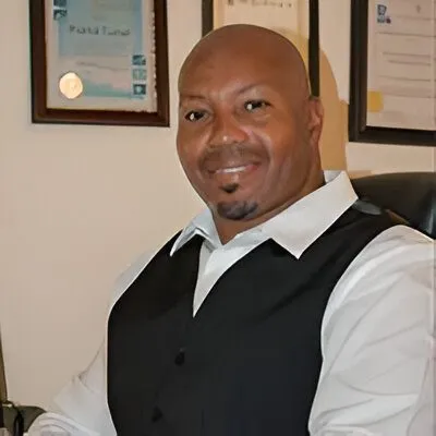 Portrait of a smiling man in a white shirt and black vest seated indoors, with framed certificates on the wall behind him.